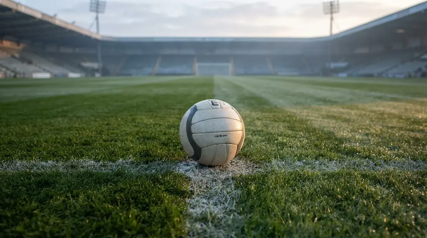 Ein einzelner Fussball liegt auf dem Anstosspunkt eines leeren Rasenplatzes am frühen Morgen, Tau glitzert auf dem Gras, die Tribüne liegt im Dunst.