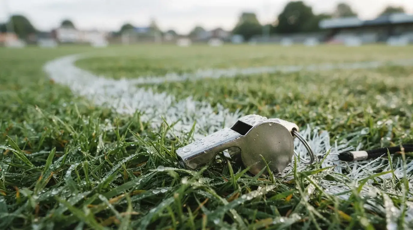 Eine Trillerpfeife liegt auf dem Rasen neben der Mittellinie, Tau hängt an den Halmen, im Hintergrund ein leerer Spielfeldrand.