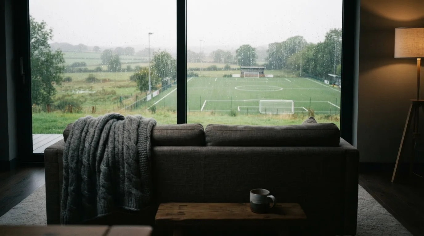 Ein Sofa mit Wolldecke steht vor einem grossen Fenster, draussen Regen und in der Ferne ein Fussballplatz.