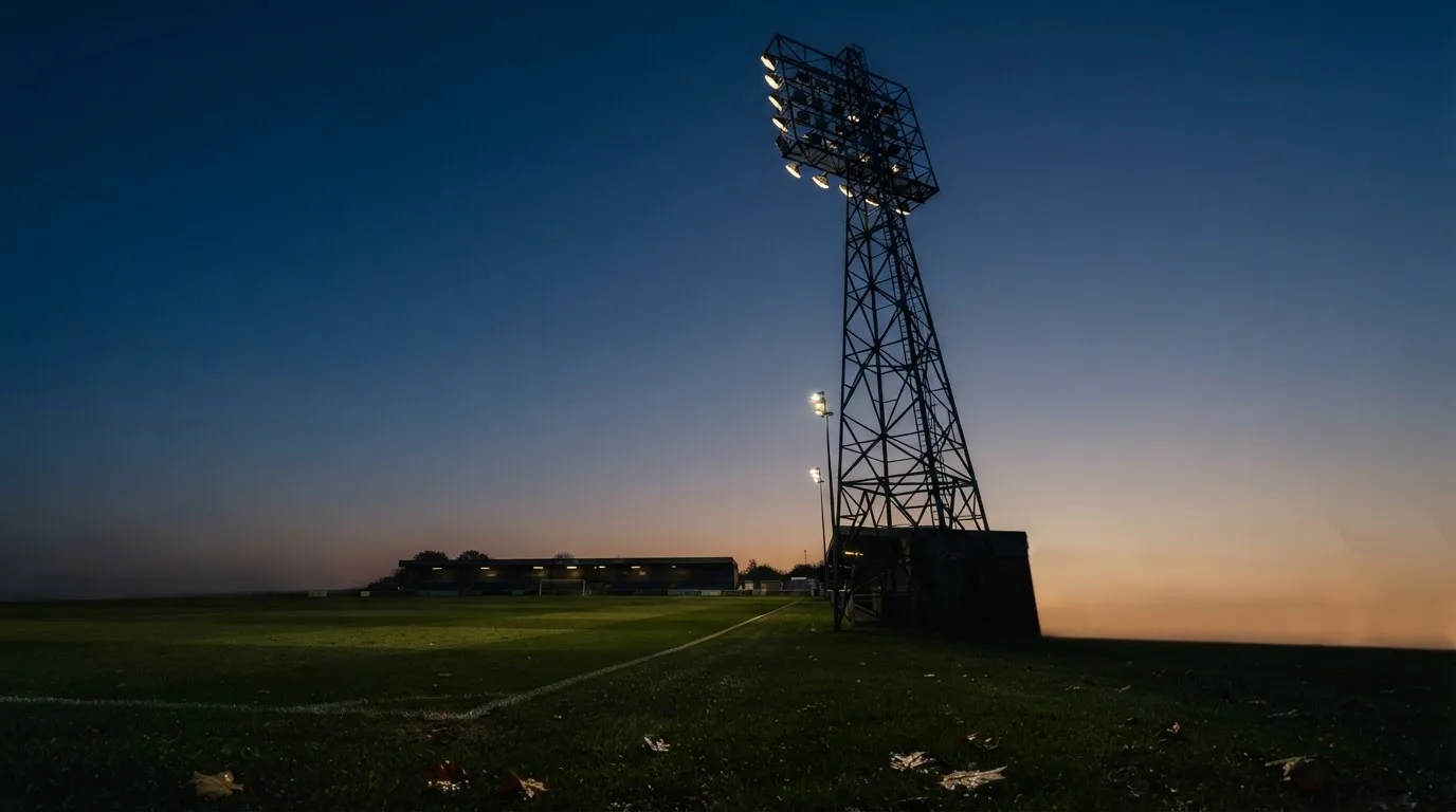 Ein Flutlichtmast ragt in den Abendhimmel, die Rasenfläche darunter liegt leer und dunkelgrün da.