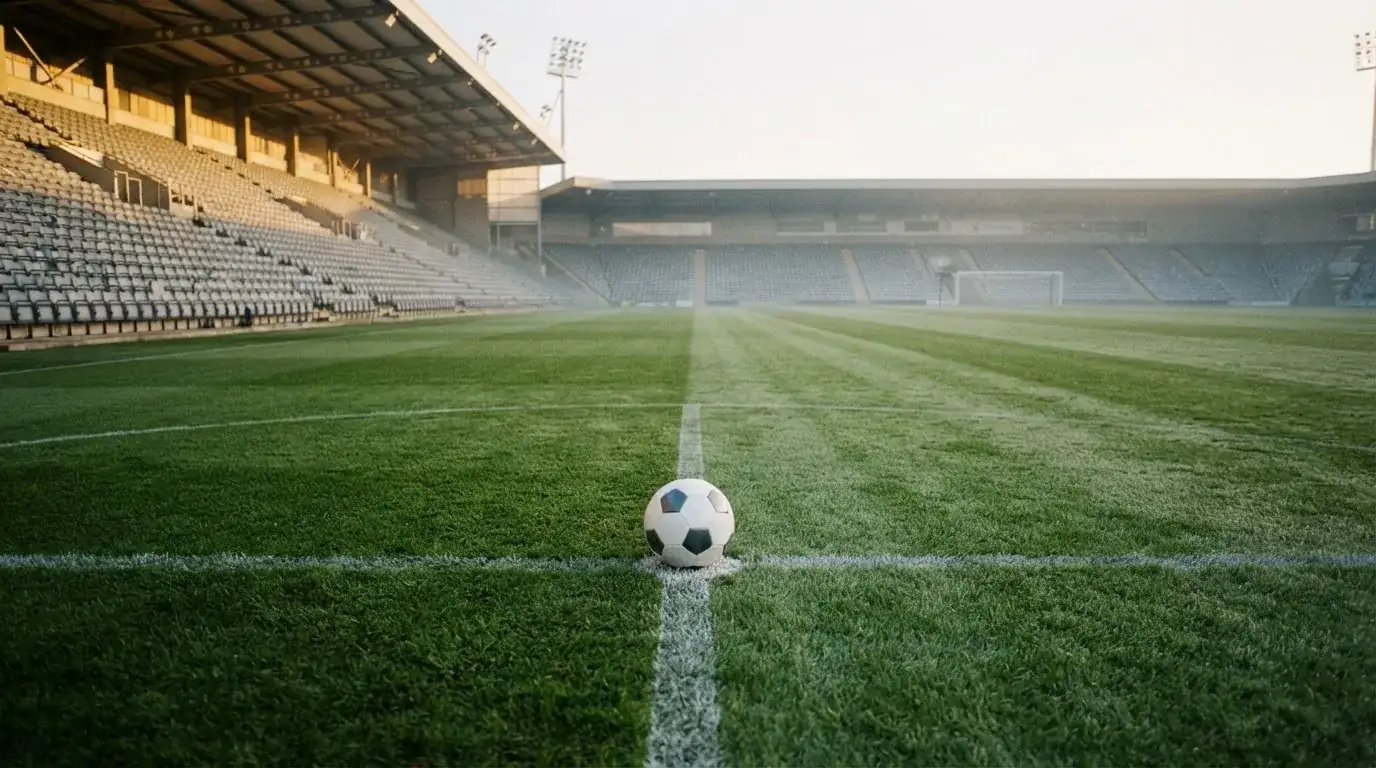 Ein Fussball liegt auf dem Anstosspunkt eines leeren Rasenplatzes im frühen Morgenlicht, dahinter erhebt sich eine menschenleere Tribüne.