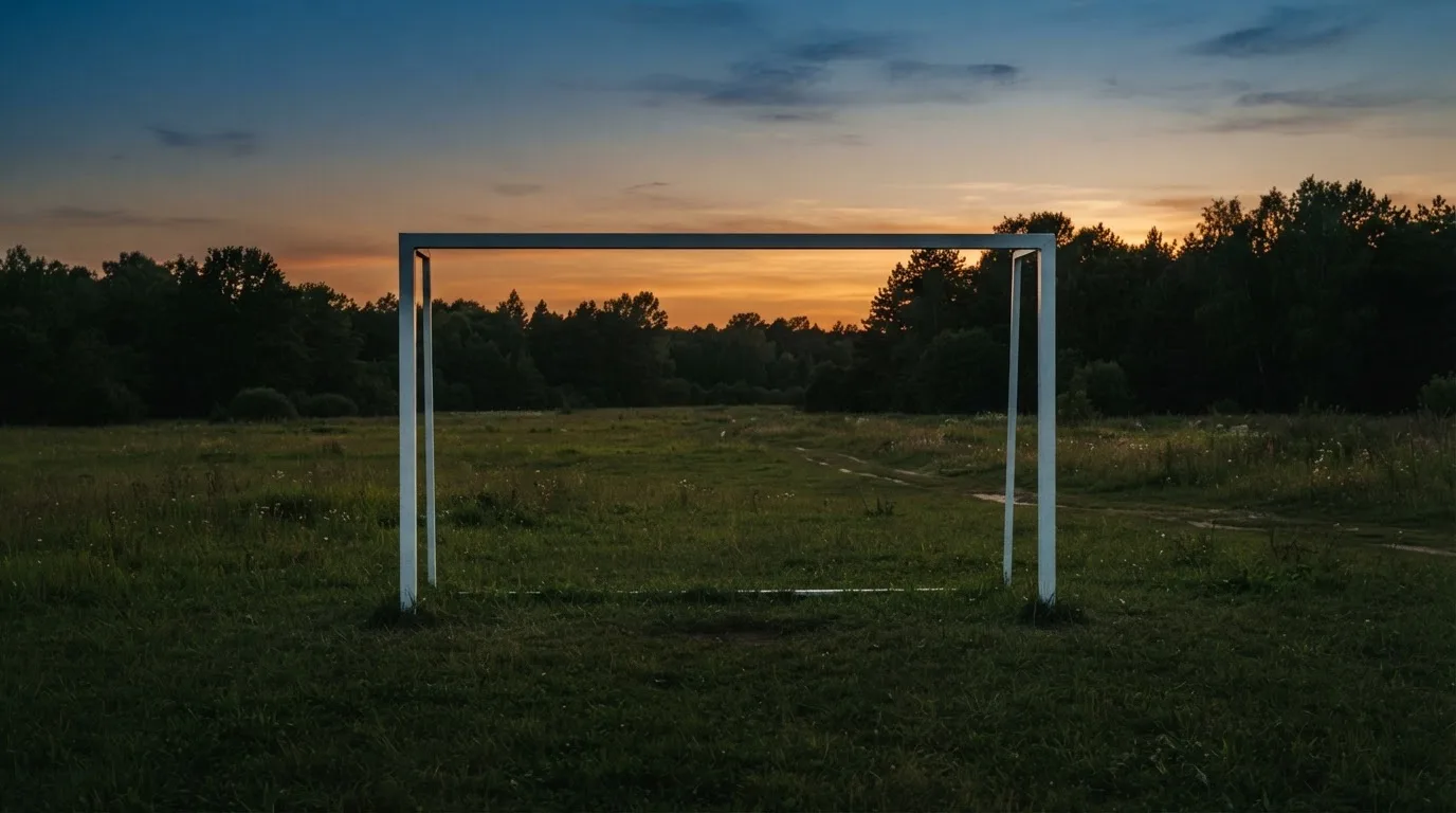 Ein Fussballtor ohne Netz steht auf einer Wiese am Waldrand, die Abenddämmerung färbt den Himmel orange.