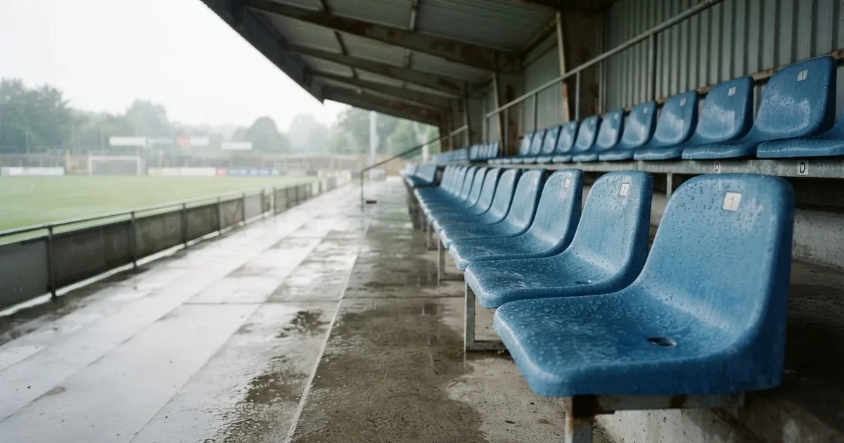 Eine Reihe leerer Sitzschalen auf der Tribüne eines kleinen Stadions, auf den Stufen glänzt Regenwasser.