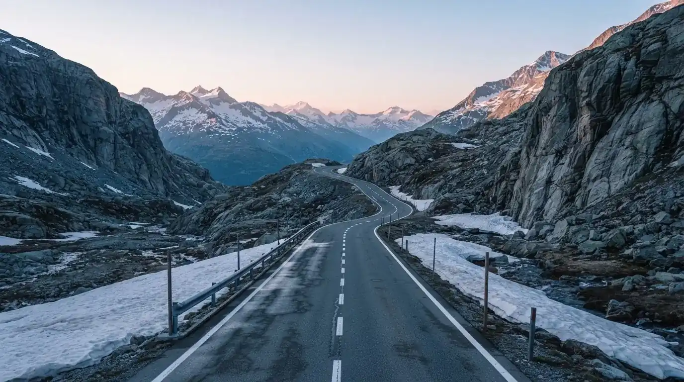 Ein Bergpass in den Schweizer Alpen mit leerer Strasse und Schneeresten am Rand im ersten Frühlicht.