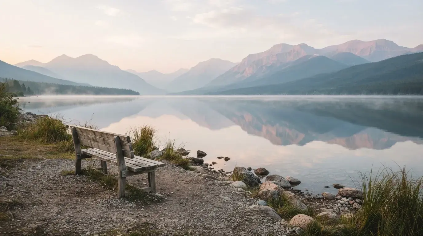 Eine Holzbank an einem Seeufer mit Blick auf eine Bergkette im Morgendunst, das Wasser ist spiegelglatt.