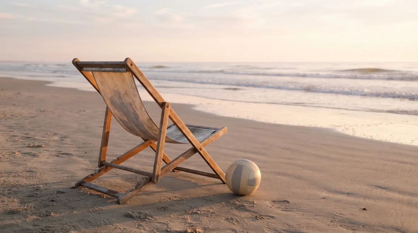 Ein leerer Liegestuhl aus Holz steht am Rand eines Sandstrands, Abendlicht und ruhige Wellen im Hintergrund.