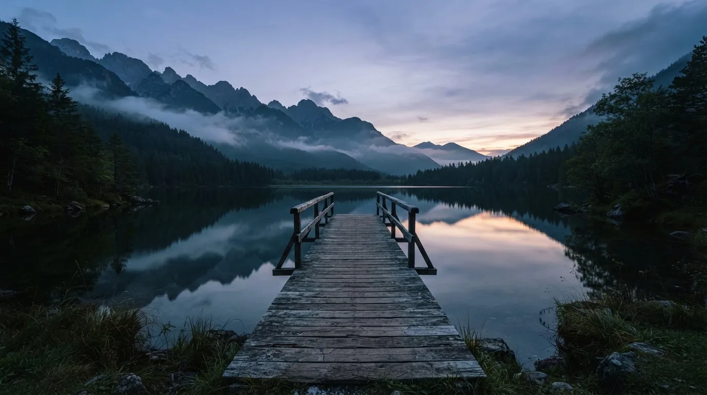 Ein Bergsee in der Dämmerung mit einem leeren Holzsteg, die Wasseroberfläche ist ruhig und Nebel zieht über die Hänge.