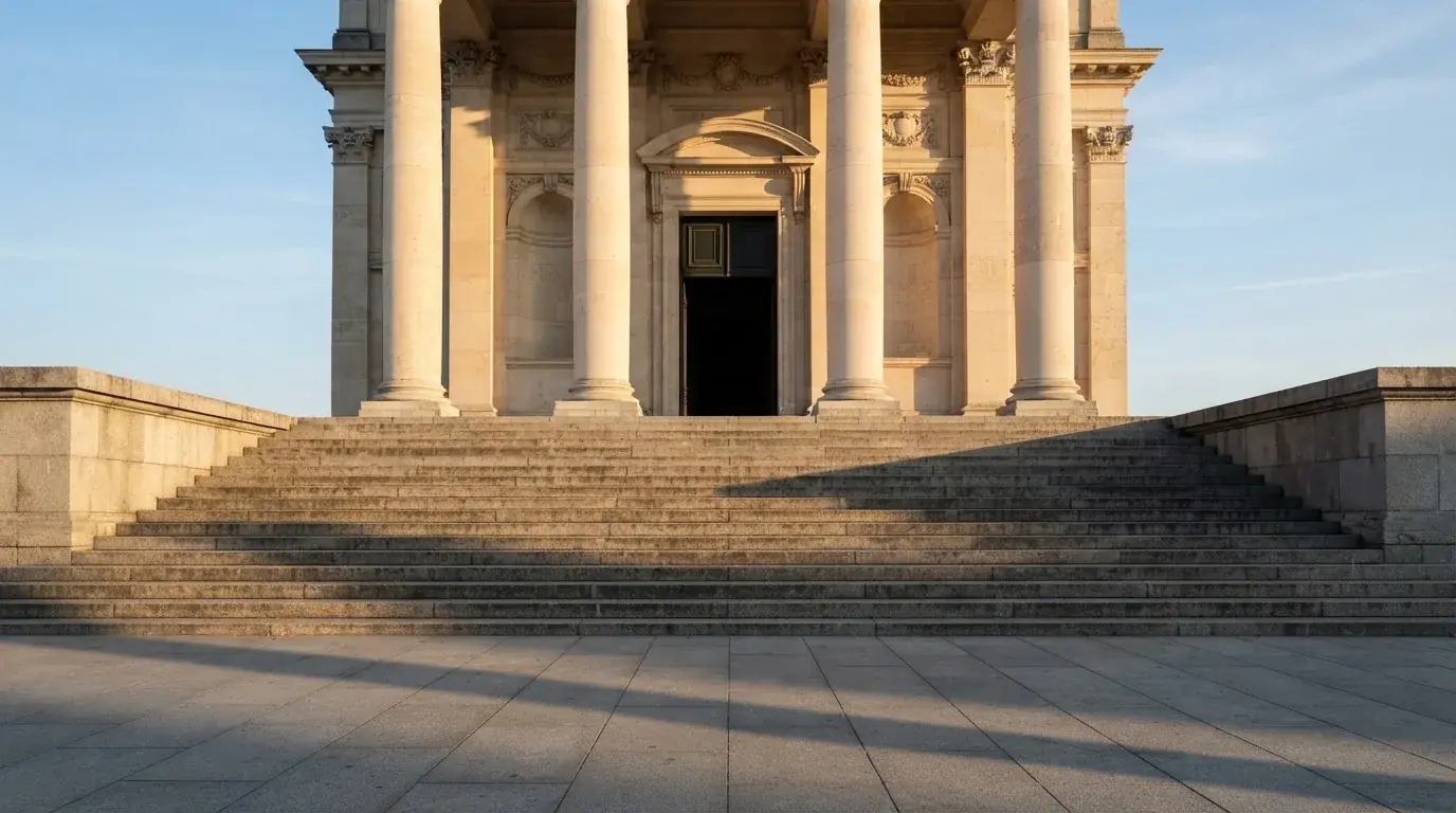 Eine steinerne Treppe führt zu einem Portal mit Säulen im Morgenlicht, der Platz davor ist leer.