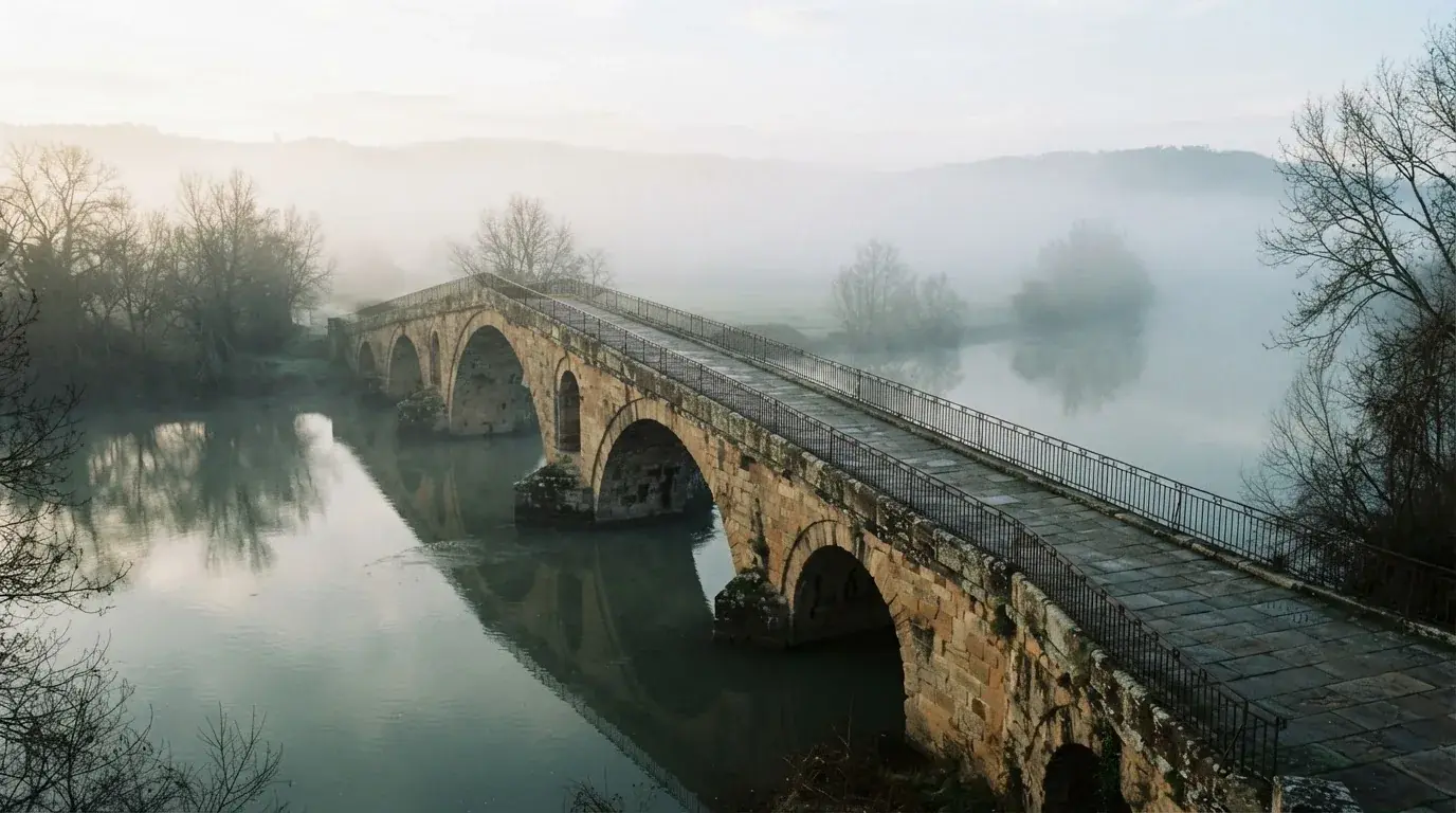 Eine Steinbrücke führt im Frühnebel über einen Fluss, auf dem Fussweg ist niemand zu sehen.